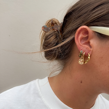A close-up side profile of a person wearing gold hoop earrings with small charms, including a green stone accent, against a plain light beige wall.