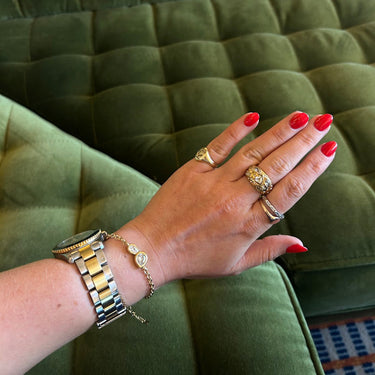 A close-up of a person's hand with red manicured nails, wearing multiple gold rings, a gold and silver watch, and a delicate gold bracelet, resting against an olive green tufted velvet sofa.